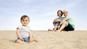 Kind am Strand schaut in die Kamera, Eltern im Hintergrund